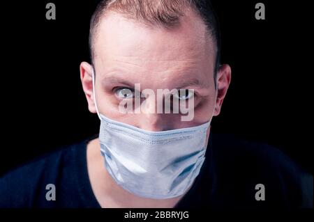 Blonder Mann mit braunen Augen mit einer medizinischen weißen Gesichtsmaske aus einer Coronavirus-Pandemie-Infektion COVID-19 isoliert auf schwarzem Backgroun Stockfoto