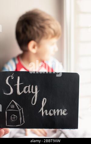 Zeichnen mit Kreide auf schwarzer Tafel, Haus mit Text Stay Home. Kind sitzt in der Nähe des Fensters auf dem Hintergrund. Konzept des Virenschutzes Stockfoto