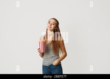 Porträt eines lachenden Mädchens in legerer Kleidung, die Popcorn Box hält und die Kamera über weißem Hintergrund isoliert betrachtet. Schöne Frau, die die Kamera betrachtet und lächelt Stockfoto