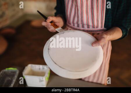 Nahaufnahme der Hände des Töpfers, die Schmuck auf keramischen Produkten machen. Platte in den Händen des Männchens. Junger Künstler Stockfoto