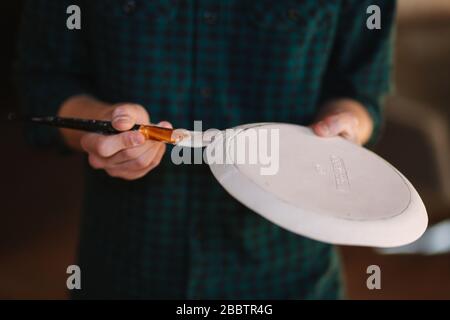 Nahaufnahme der Hände des Töpfers, die Schmuck auf keramischen Produkten machen. Platte in den Händen des Männchens. Junger Künstler Stockfoto