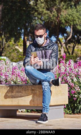 Mann mit Maske sitzt im Freien auf dem Smartphone. Selbstvorkehrungen während einer Pandemie von Coronavirus. Stockfoto