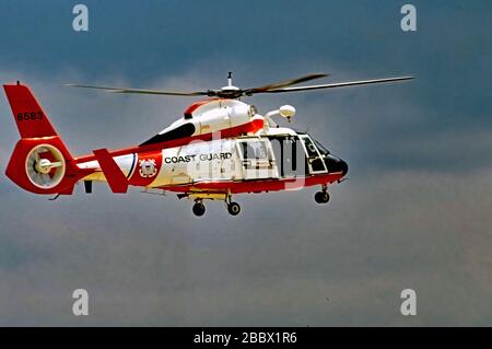 Camp Springs Maryland, USA, 20. Mai 1989 EIN Hubschrauber der United States Coast Guard MH-65 Dauphin Short Range Recovery wechselt in den Hover-Modus, während der Rettungsschwimmer im Rücken sich darauf vorbereitet, den Hebegeschirr im Rahmen einer Demonstration der Fähigkeiten des von Airbus hergestellten Hubschraubers einzusetzen, den die Küstenwache für verschiedene Missionen verwendet, einschließlich Jetzt mit Maschinengewehrversion (MH-65CS) bewaffnet, um das US-Kapitol zu schützen Stockfoto