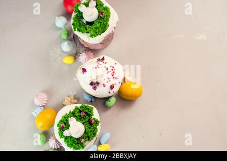 Mit Sahne verzierte orthodoxe Osterkuchen in Form von Gras- und Kaninchenfigürchen aus weißer Schokolade auf grauem Grund neben bemalten Eiern. Easter Food Konzept. Idee für Kinder. Stockfoto