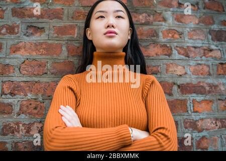 Nahaufnahme der jungen schönen asiatischen Frau, die selbstbewusst aussieht und vor Backstein-Wandhintergrund steht. Stockfoto