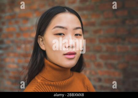 Nahaufnahme der jungen schönen asiatischen Frau, die selbstbewusst aussieht und vor Backstein-Wandhintergrund steht. Stockfoto