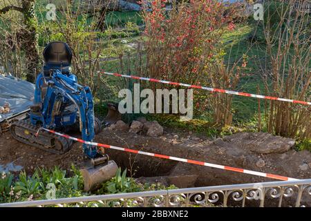 Blauer Bagger zwischen provisorischer Bandsperre im Hinterhof. Rot-weiße Barrikade für die Baustelle des Abwasserschaches. Stockfoto