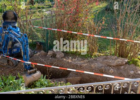 Blauer Bagger zwischen provisorischer Bandsperre im Hinterhof. Rot-weiße Barrikade für die Baustelle des Abwasserschaches. Stockfoto