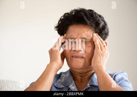 Asiatische alte Frau, die auf dem Sofa sitzt und zu Hause Kopfschmerzen hat. Konzept für das Gesundheitswesen. Stockfoto