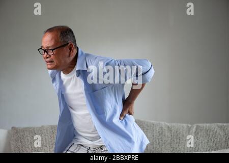 Asiatischer alter Mann sitzt auf dem Sofa und hat Rückenschmerzen, Rückenschmerzen zu Hause. Konzept für das Gesundheitswesen. Stockfoto