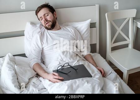 Der Mann war sehr müde und schlief im Bett mit einem Ultrabook ein, nachdem er seine Arbeit aus der Ferne beendet hatte. Quarantänekonzept. Stockfoto