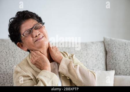 Asiatische alte Frau, die auf dem Sofa sitzt und Nackenschmerzen hat, Rückenschmerzen zu Hause. Konzept für das Gesundheitswesen. Stockfoto