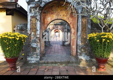 Uralter Korridor mit verwitterten Backsteinwänden und fernen Portalen mit gelben Blumen in einem Blumentopf. Hue Imperial Palace Verbotene Stadt, Vietnam Asien Stockfoto