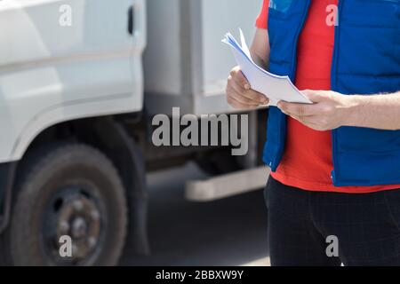 Kurierfahrer in Uniform macht Notizen in Dokument und Lieferung weißer LKW behinde ihn Stockfoto