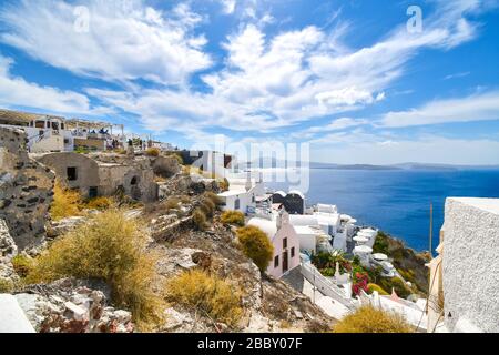 Blick vom Dorf Oia auf der Bergkuppe über die Caldera und das blaue Meer mit einer kleinen Kirche, einem Resort und alten Gebäuden. Stockfoto