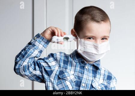 Der junge Junge mit schmerzenden Augen in einer medizinischen Maske misst die Temperatur eines Thermometers im Ohr. Medizinischer Schutz vor durch die Luft übertragenen Krankheiten, Koronav Stockfoto