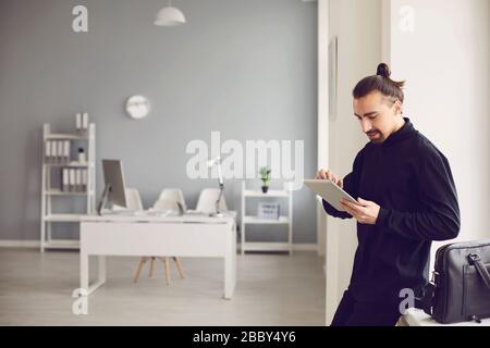 Ein ernster Kerl steht mit einem Tablett in der Hand, während er im Büro am Arbeitsplatz steht. Stockfoto