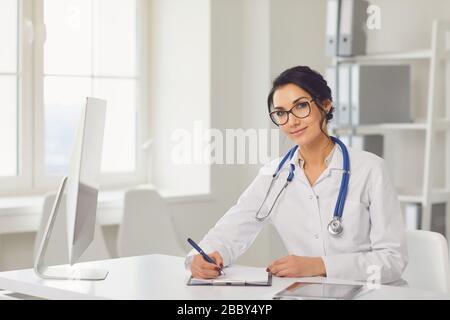 Selbstbewusste Frau Ärztin Kinderärztin schreibt in einer Zwischenablage, die an einem Tisch in einem weißen Büro des Krankenhauses sitzt Stockfoto