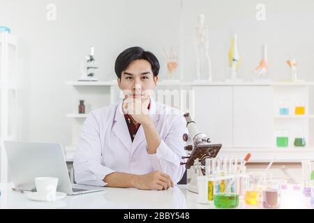 Junger, gutaussehender Wissenschaftler, der im Laborarbeitstisch sitzt, Asian Race, der mit der Kamera Hand auf das Kinn blickt Stockfoto