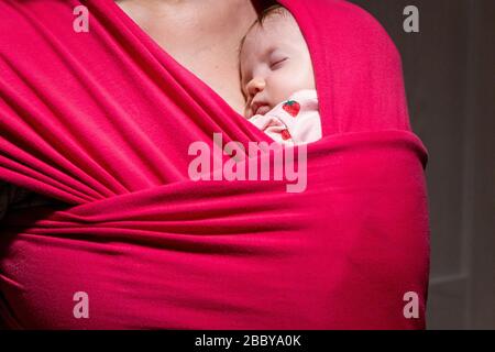 Mutter, die ihre niedliche Tochter in Schlinge trägt. Neugeborenes Baby, das in einer Schlinge schläft, in der Umarmung ihrer Mutter. Das Mädchen ist 2 Monate alt. Stockfoto