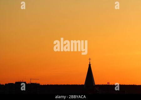 Sonne unter dem Horizont und das Dach der Kirche mit einem Kreuz auf dem Hintergrund feuriger dramatischer orangefarbener Himmel bei Sonnenuntergang oder Sonnenaufgang, der von der Sonne hinterleuchtet wird. Platz für Text und Design. Stockfoto