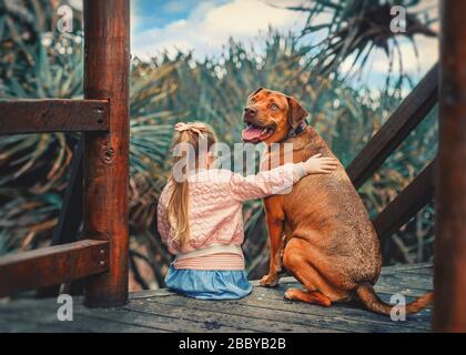 Süßes kleines Mädchen umarmt einen großen roten ungarischen vizsla-hund, der zusammen auf der Holztreppe sitzt. Kind und Haustier. Stockfoto