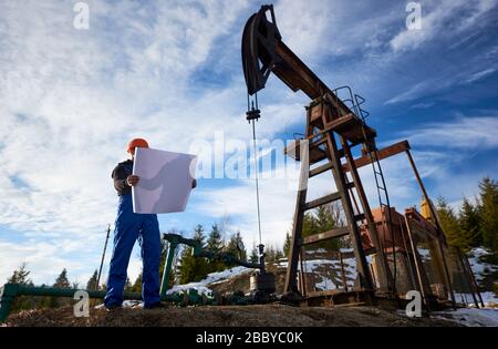 Mann in blauer Uniform und orangefarbener Helm im Ölfeld stehend mit bedrucktem Plan, mit überdachtem Gesicht, neben dem Ölpumpenheber an schönem sonnigen Tag, Blick in den niedrigen Winkel. Konzept der Öl- und Gasindustrie Stockfoto