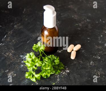 Braune Glasflasche mit ätherischem Öl oder Creme, Vitaminen und heilenden grünen Kräutern auf dem dunkel rauhen Texturhintergrund. Stockfoto