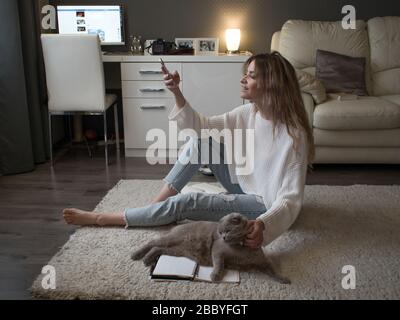 Planen Sie Dinge, setzen Sie Ziele. Eine junge Frau sitzt auf dem Boden auf dem Teppich und schreibt in ein Tagebuch. Behalten Sie einen Gewohnheits-Tracker und verwenden Sie die App auf dem Smartphone. Stockfoto