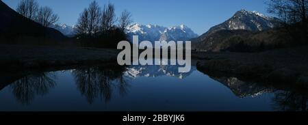 Farchant, Deutschland. April 2020. Die schneebedeckten Gipfel des Wettersteingebirges mit Alpspitze (M) und Zugspitze (2. Von rechts) spiegeln sich zusammen mit dem Kramerspitz (Ammergauer Alpen, r) in einem Teich wider. Credit: Angelika Warmuth / dpa / Alamy Live News Stockfoto