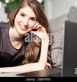 Hübsche junge Frau, die mit einem Laptop auf der Couch liegt und sympathisch an der Kamera lächelt Stockfoto