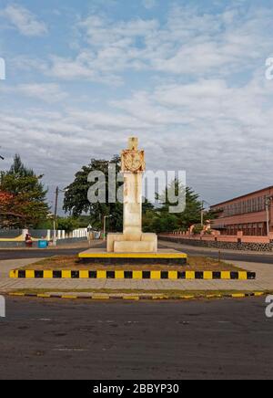 Ein Denkmal im sowjetischen Stil in Sao Tome im Zentrum eines kleinen Kreisverkehrs mit einer von Bäumen gesäumten Allee dahinter. Stockfoto