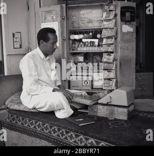 1960er, historisch, Geldwechsel auf der Straße, ein Mann, der auf einem Kissen auf einem Teppich sitzt und Bankscheine zählt, Jeddah, Saudi-Arabien. Münzen und Banknoten sind in einem offenen Safe neben ihm verstaut. Ein Facit Calculator der Ära mit Griff befindet sich vor ihm auf dem Teppich. Stockfoto