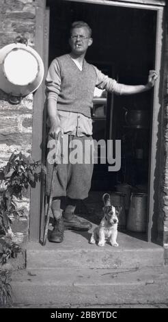 Dreißiger Jahre, ein Landarbeiter in der Kleidung, die für die Epoche typisch ist, einschließlich Mehl und Zigarette im Mund, mit Haustierhund, der in der Tür seines Cottages, Devon, England, steht. Eine Reihe von Milchfässern ist im Eingang zu sehen, während außen an der Wand neben der Tür eine Melkschale aus Metall hängt. Stockfoto