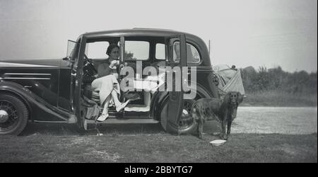 1930-40s, historisch, eine Dame sitzt in einem 4-Türer Salon-Motorwagen der Ära - vielleicht ein Riley Adelphia, da es 6 Seitenfenster, möglicherweise ein Fiat 518 oder Ardita - auf einem Grasrand geparkt mit einem Picknick, mit ihrem Haustier Hund, ein Retreiver für Unternehmen. Beachten Sie die Leinwand aussehende Packung auf der Rückseite des Autos und die hintere Tür an der Rückseite anstatt der Vorderseite, manchmal auch als "Bustür" bekannt. Meist auf 4-Türer-Limousinen oder Limousinen beschränkt, hatten sie Vorteile, nämlich das Fahrzeug leichter zu machen und auszusteigen, was zum Beispiel Chauffern einen leichteren Zugang zur Hecktür ermöglicht. Stockfoto