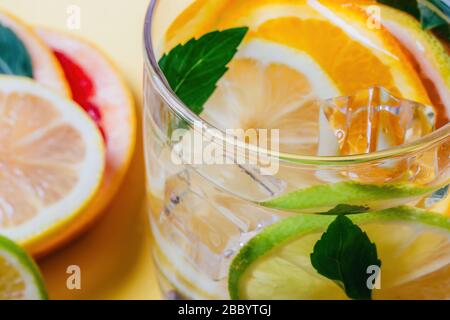 Zitrusfrüchte-, Limetten-, Orange- und Grapefruitscheiben mit Minzblättern und Eiswürfeln in einem Glas Wasser und in Haufen auf gelbem Hintergrund. Nahaufnahme. Stockfoto