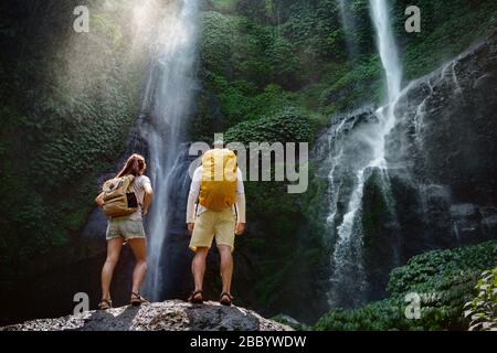 Ein paar Reisende oder Wanderer mit Rucksäcken stehen auf einem großen Felsen und genießen einen großen Wasserfall Stockfoto
