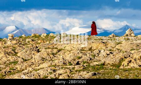 Buddhistischer Pilger in roter Robe, der am Nam Tso umherwandert. Für tibeter ist das Gebiet um den See heilig. Mit Wolken und schneebedeckten Bergen. Stockfoto