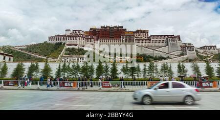 Vorderansicht (Panorama) des Potala Palastes. Ein silberfarbenes Auto kommt von rechts nach links vorbei. Fußgänger gehen vor dem Palast. Stockfoto