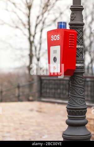 Hilfe- oder Polizeiruftaste. SOS. Auf der Straße, auf einem Lampenpfosten, ist ein Ständer mit einem Knopf befestigt. Rotes Gehäuse mit blauer Warnleuchte oben. Stockfoto