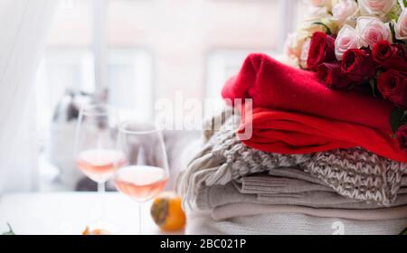 Ein Stapel Strickpullover und ein Blumenstrauß aus roten und weißen Rosen in der Nähe des Fensters. Homeless, warme Kleidung, eine schöne Katze und zwei Gläser Wein.das Konzept der Geschenke und Überraschungen.Banner.Soft Focus Stockfoto