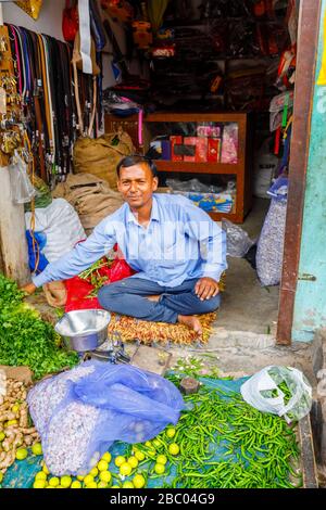 Ladenbesitzer, der in der Tür eines kleinen Geschäftes sitzt, das Gemüse verkauft, Straßenszene im Distrikt Mahipalpur, einem Vorort in Neu-Delhi, Indien Stockfoto