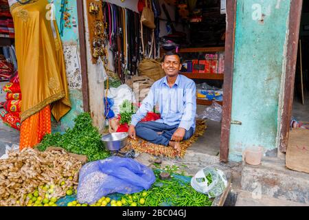 Ladenbesitzer, der in der Tür eines kleinen Geschäftes sitzt, das Gemüse verkauft, Straßenszene im Distrikt Mahipalpur, einem Vorort in Neu-Delhi, Indien Stockfoto