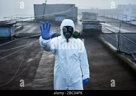 Forscher, der auf dem Dach eines Hochhauses steht und weißen Schutzanzug, blaue Handschuhe und Gasmaske trägt, mit Blick auf die Kamera, gestiertes Stoppschild Stockfoto