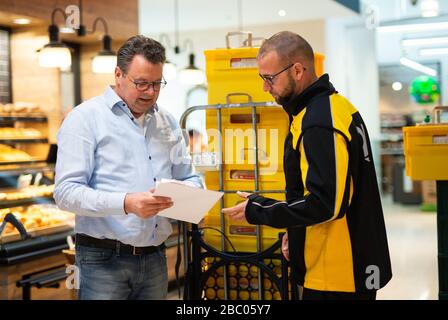 Gangelt Birgden, Deutschland. April 2020. Heinz Schmiz, Rewe-Geschäftsführer, übergibt Lebensmittel an Michael Peltzer (r), Postbote in Gangelt, im Rewe-Supermarkt Schmiz. Im Kreis Heinsberg, der besonders vom Corona-Virus betroffen ist, startet die Deutsche Post ein Pilotprojekt zur Bereitstellung grundlegender Dienstleistungen für Risikogruppen, die von der Post mit Lebensmitteln und Haushaltswaren aus dem lokalen Einzelhandel versorgt werden können. Credit: Jonas-Güttler / dpa / Alamy Live News Stockfoto