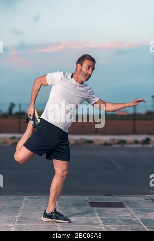 Kaukasischer Athlet man in weißem T-Shirt, streckt die Beinmuskeln im Gleichgewicht, an einem Tag mit blauem Wolkenhimmel bei Sonnenuntergang. Stockfoto