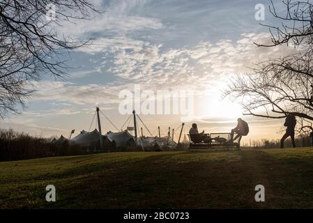 Frühlingsstimmung an einem milden Februarabend im Münchner Olympiapark. Im Vordergrund Menschen auf einer Bank, im Hintergrund das Zeltdach des Olympiastadions. [Automatisierte Übersetzung] Stockfoto