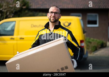 Gangelt Birgden, Deutschland. April 2020. Michael Peltzer, Postbote in Gangelt, steht mit einem Paket vor einem Postbus. Im Kreis Heinsberg, der besonders vom Coronavirus betroffen ist, startet die Deutsche Post ein Pilotprojekt zur Bereitstellung von Grundleistungen für Risikogruppen, die von der Deutschen Post mit Lebensmitteln und Haushaltswaren des örtlichen Einzelhandels versorgt werden können. Credit: Jonas-Güttler / dpa / Alamy Live News Stockfoto