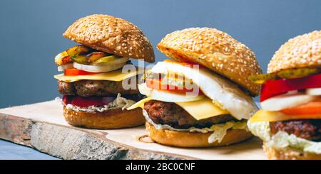3 saftige Burger auf Baumrinde. Verschiedene Käsebrenner auf grauem Hintergrund. Stockfoto