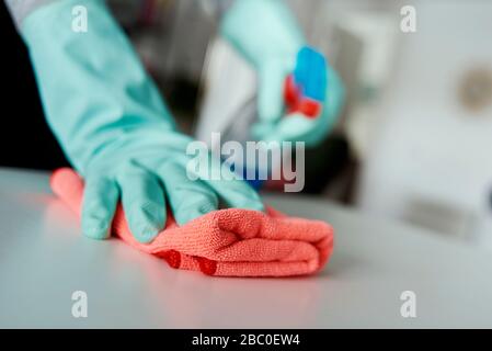 Nahaufnahme eines Mannes, tragen von Gummihandschuhen, Reinigen der Oberfläche eines weißen Tisches mit einem Geschirrtuch und einem Reinigungsmittel aus einer Sprühflasche Stockfoto